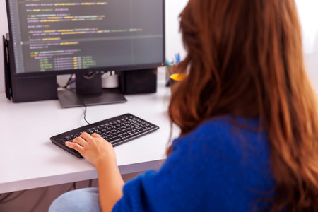 Female programmer coding while working remotely from home; female web developer working in home office using desktop computer. Focus on hand on keyboardの写真素材