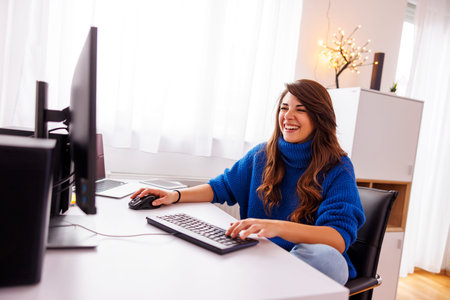 Female freelancer working in home office sitting at her desk, using desktop and laptop computers;...