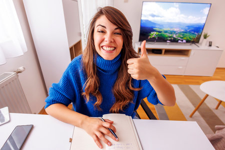 Young woman showing thumbs during online meeting while working remotely from home; woman working in home office having conference callの写真素材