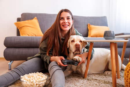 Young woman having fun spending leisure time at home with her pet, beautiful Labrador dog, playing video games and eating popcornの写真素材