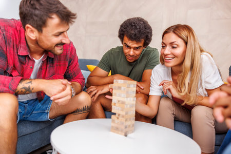 Group of cheerful young friends having fun playing block stacking and removing balance game while spending leisure time together at homeの写真素材