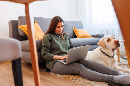 Beautiful young woman sitting on living room floor with with her dog lying beside her working remotely from home using laptop computerの写真素材