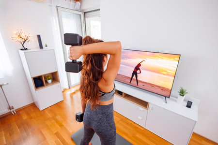 Active young woman in sportswear doing arm workout using dumbbells at homeの写真素材