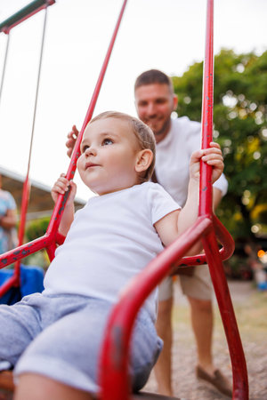 Young father having fun playing with his son on the playground in the park, swinging him in a swingの写真素材
