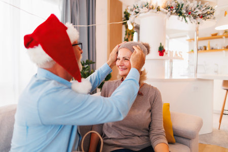 Senior couple sitting on the sofa celebrating Christmas at home, having fun while exchanging and opening presents, man putting hat on woman's headの写真素材