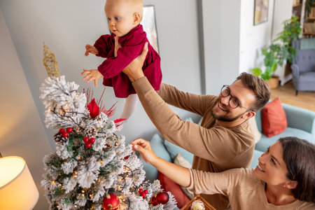 High angle view of young parents having fun decorating Christmas tree with their cute little baby girl, father lifting daughter to place tree topper starの写真素材
