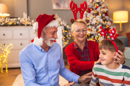 Grandparents having fun playing with grandson at home on Christmas day, putting on Santa hat and costume reindeer antlersの写真素材