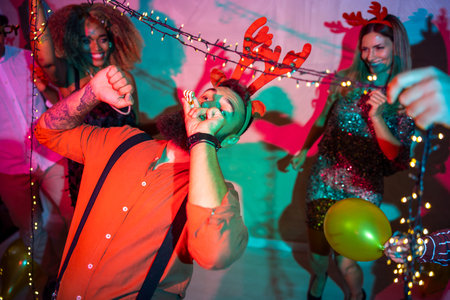 Group of cheerful young friends having fun doing limbo dance passing under Christmas lights at New...