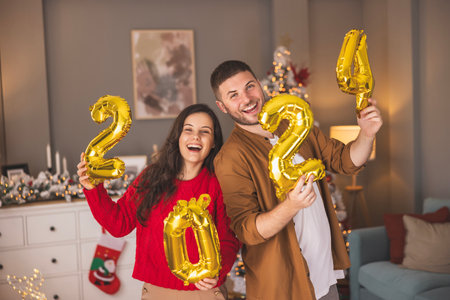 Beautiful young couple in love having fun decorating home for winter holiday season, holding balloons shaped as numbers 2024 representing the upcoming New Yearの写真素材