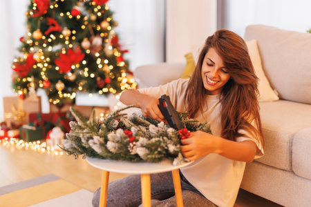 Beautiful young woman having fun decorating home for Christmas, making a Christmas wreath attaching decorations using glue gunの写真素材