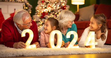 Grandparents celebrating New Years Eve at home with kids, sitting by the Christmas tree, holding illuminative numbers 2024 representing the upcoming New Yearの写真素材