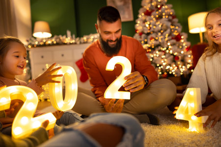 Young parents and children having fun celebrating New Year at home, holding illuminative numbers 2024 while sitting by the Christmas treeの写真素材