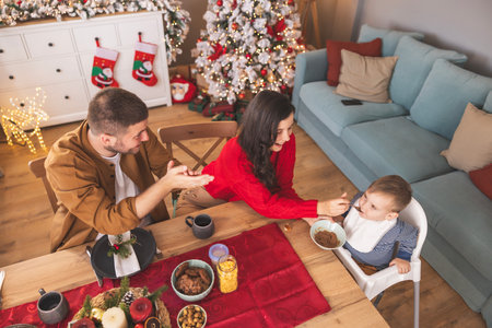 Father drinking coffee and mother feeding her cute little baby boy while having Christmas morning breakfast at homeの写真素材