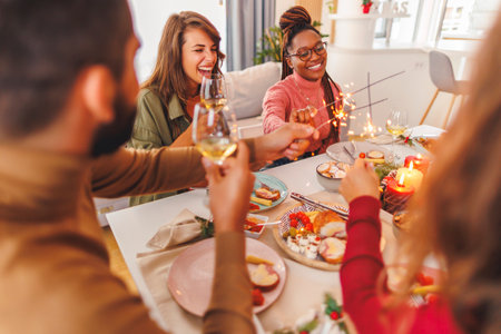 Group of cheerful young friends having Christmas dinner together at home, waving with sparklers and making a toast raising glasses of wineの写真素材