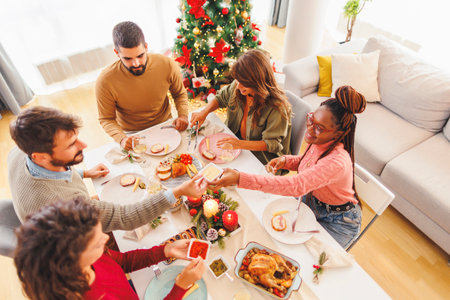 High angle view of group of cheerful young friends having Christmas dinner together at home, passing the food dishes to each other and having conversationの写真素材