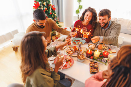 Group of cheerful young friends having Christmas dinner together at home, waving with sparklers and making a toast raising glasses of wineの写真素材