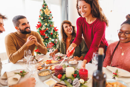 Group of cheerful friends celebrating Christmas gathered at home, woman lighting table centerpiece candle while hosting Christmas dinner party for friendsの写真素材
