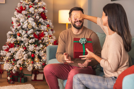Happy couple in love exchanging Christmas presents while spending Christmas day together at home, wife surprising husband holding his eyes closedの写真素材