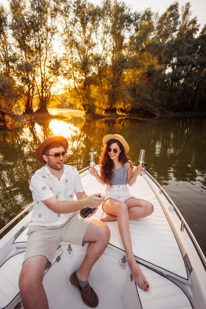 Beauitful young couple in love enjoying summer vacation, opening a bottle of champagne celebrating anniversary while sailing to the sunset on a boatの写真素材