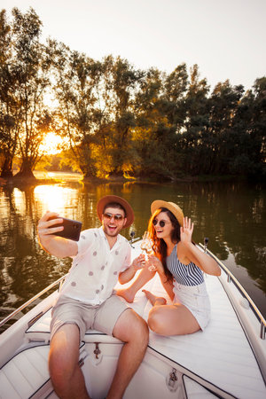 Beauitful young couple in love having fun on summer vacation, taking a selfie and making a toast raising glasses of champagne while sailing to the sunset on a boatの写真素材