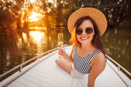 Beautiful young woman relaxing while on summer vacation, drinking champagne and sailing to the sunset on a boatの写真素材