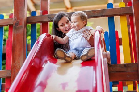 Beautiful young mother and cute baby boy having fun playing on a slide in the park, mother helping toddler son to climb on the topの写真素材