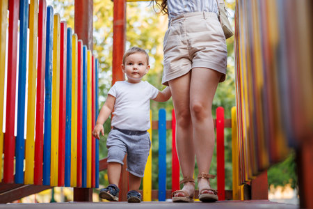 Beautiful mother and toddler son having fun taking a walk and playing on the playground in the park, climbing on a tree houseの写真素材