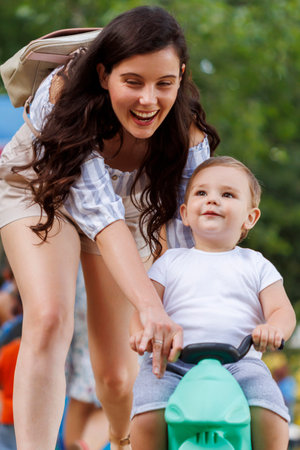 Cheerful young mother enjoying leisure time outdoors with her cute little toddler son, teaching him how to ride a bikeの写真素材