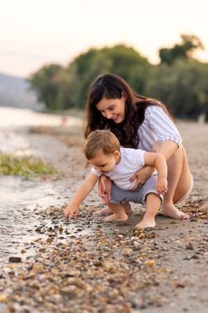Beautiful young mother having fun playing with her toddler son while spending summer afternoon on the beach together, hugging him and throwing pebbles in waterの写真素材