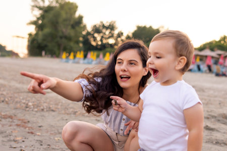 Beautiful young mother having fun playing with her baby boy while spending summer afternoon on the beach together, hugging him and throwing pebbles in waterの写真素材