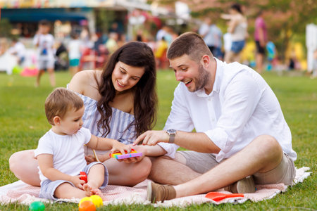 Beautiful young parents having picnic in the park with their cute little baby boy, sitting on the blanket and playing with colorful ballsの写真素材