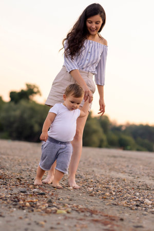 Beautiful young mother walking down the beach with her cute little baby boy holding his hand and enjoying leisure time outdoors togetherの写真素材