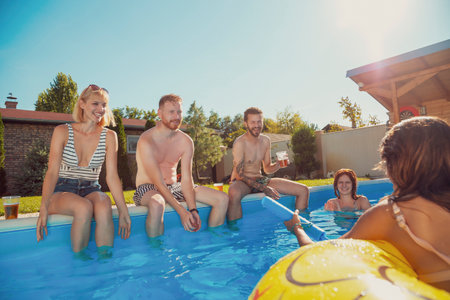 Group of cheerful young friends having fun at an outdoor summertime party, sitting by the pool, drinking beer and splashing water on each otherの写真素材