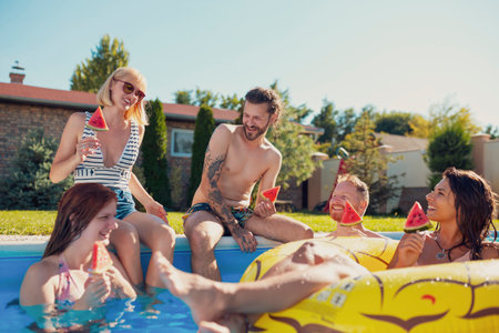 Group of young friends having fun at summertime swimming pool party, sitting by the pool and eating watermelon popsicles, spending sunny summer day outdoorsの写真素材