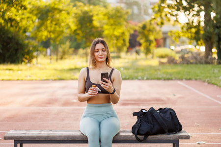 Active young woman in sportswear sitting on the bench by the basketball court eating an energy bar and using smart phone while taking a workout breakの写真素材