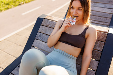 Active young woman in sportswear sitting on the bench outdoors eating a protein bar while taking a workout breakの写真素材