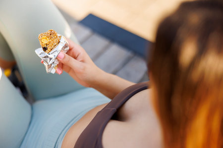 Active young woman in sportswear sitting on the bench outdoors eating a protein bar while taking a workout breakの写真素材
