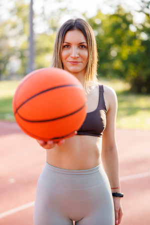 Active young woman in sportswear having fun playing basket at an outdoor basketball court, holding ball and showing it towards the cameraの写真素材