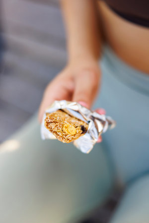 Detail of active fit woman in sportswear sitting on the bench holding a protein bar while taking a workout breakの写真素材