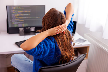Female software developer stretching out while sitting and working at her desk in home office;...