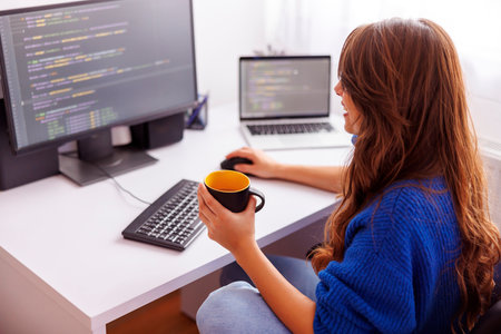 Female programmer drinking coffee while working remotely from home; female software developer having a cup of coffee while working in home officeの写真素材