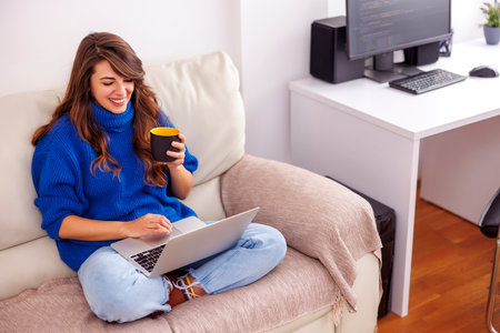 Young woman sitting on the living room couch, holding laptop computer in her lap and taking a coffee break while working from homeの写真素材