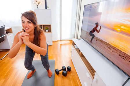 Active young woman doing online yoga session at home, following exercises shown by instructor on TVの写真素材