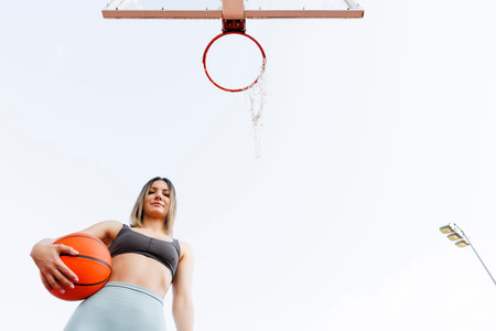 Low angle view of young woman in sportswear holding a ball taking a break while playing basketball as morning workout routineの写真素材