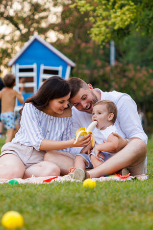 Cute little baby boy sitting in his fathers lap and eating a banana while on a picnic with his parents in the parkの写真素材