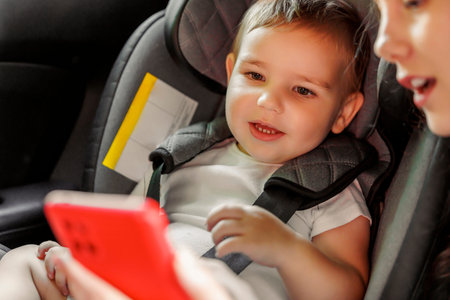 Mother and toddler son having fun while riding on back seat of the car, mother playing songs and cartoons to son using smart phone while baby is safely placed in car seatの写真素材
