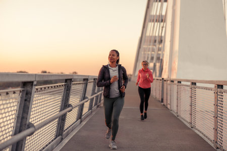 Fit active women in sportswear jogging over the bridge at dawn, enjoying beautiful sunrise while doing outdoor morning workout routineの写真素材