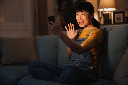 Beautiful woman relaxing at home late at night, having video call using smart phone, speaking and wavingの写真素材