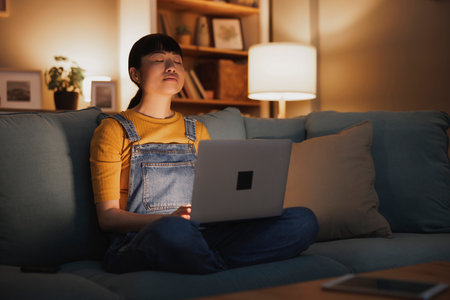 Beautiful woman sitting on living room couch meditating with laptop computer in her lap while working remotely from home late at nightの写真素材