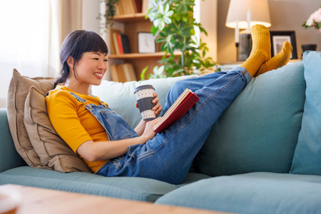 Beautiful woman sitting on living room couch reading a book and drinking coffee while relaxing at homeの写真素材
